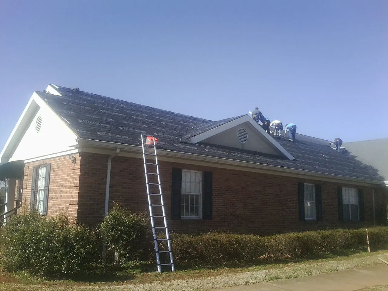 Crew tearing shingles off a brick ranch roof with a ladder set — Metal Roof Repair work by our crew in Saint Anne