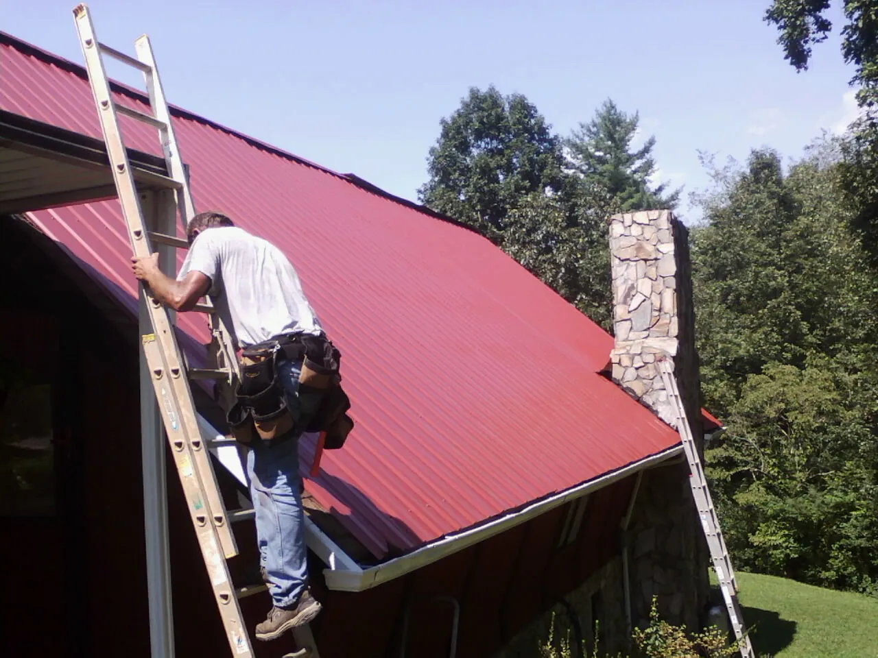 Roofer on a ladder beside a red metal roof and stone chimney — Soffit & Fascia Repair by Roofing Brothers in Saint Anne