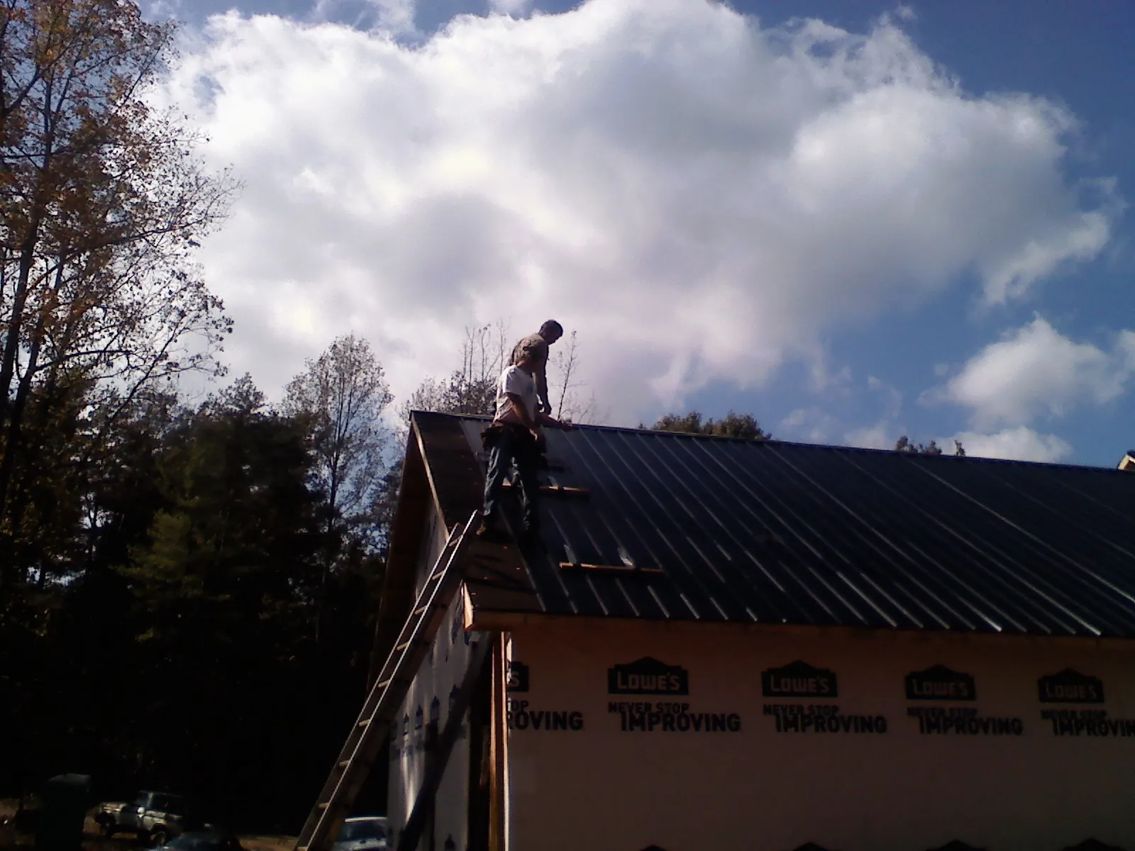 Roofer fastening dark metal panels high against a cloudy sky in Saint Anne